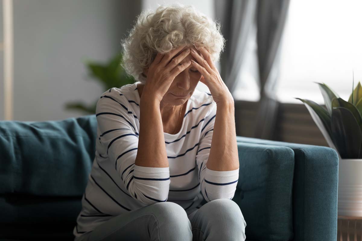 A woman sitting on a couch, resting her hands on her head, appears deep in thought or stressed.