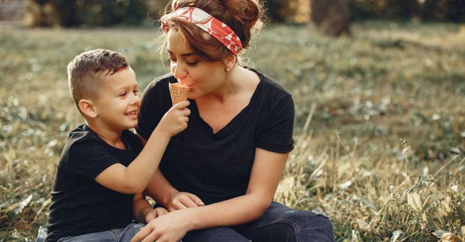 A woman and a child enjoy ice cream together while sitting on the grass, surrounded by a sunny outdoor setting.