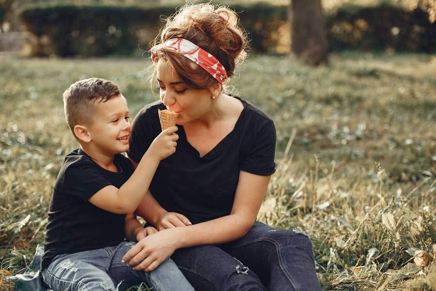 A woman and a child enjoy ice cream together while sitting on the grass, surrounded by a sunny outdoor setting.